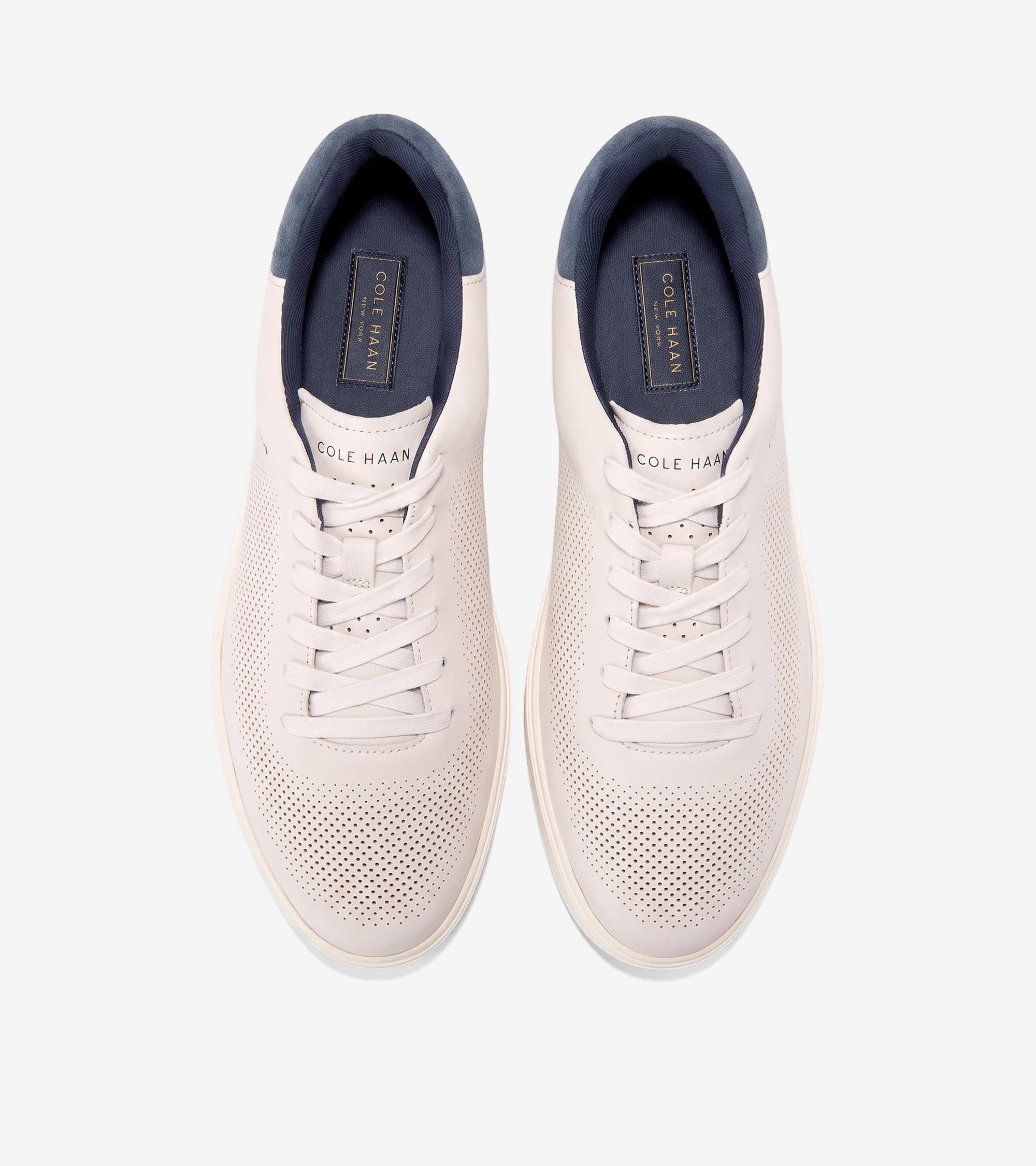 Pair of white Cole Haan GP Angleace sneakers with perforated leather uppers and navy accents, seen from a top-down view.